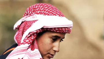 در این تصویر، یک فرد با چهره‌ای متعلق به سن جوانی دیده می‌شود. او یک کلاهی با طرح قرمز و سفید بر سر دارد که به شیوه‌ای سنتی پوشیده شده است. چهره‌اش به سمت پایین متمایل است و نگاهی تفکرآمیز دارد. پس‌زمینه تصویر مبهم است و توجه بیشتر بر روی شخص متمرکز شده است. این تصویر حس آرامش و تمرکز را منتقل می‌کند.