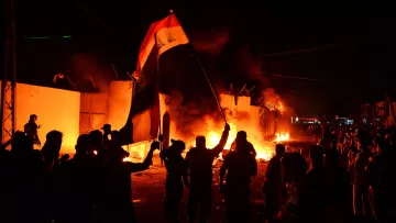 L'image montre une scène nocturne animée, probablement lors d'une manifestation ou d'une émeute. Des personnes brandissent un drapeau, en l'occurrence le drapeau irakien, tandis que des flammes s'élevées en arrière-plan, suggérant des tensions ou des troubles. Les silhouettes des manifestants sont bien visibles, et l'ambiance est chargée d'énergie et d'émotion. La lumière des feux crée un contraste fort avec l'obscurité de la nuit.