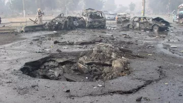 L'image montre un site de dévastation après une explosion. On peut voir plusieurs voitures endommagées et partiellement détruites, gisant sur le sol. Au centre, un cratère profond et large est présent dans le bitume, indiquant la force de l'explosion. L'environnement semble chaotique avec des débris éparpillés et une atmosphère troublée, probablement à cause de fumée ou de poussière. Cette scène évoque une situation d'urgence ou un acte de violence, reflétant des dommages importants.