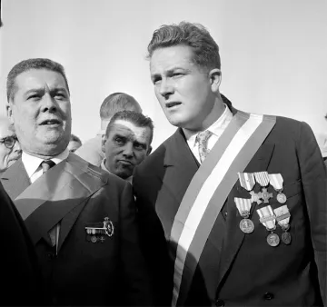 L'image montre deux hommes qui semblent participer à un événement officiel ou une cérémonie. L'un des hommes porte plusieurs médailles accrochées à sa veste, ainsi qu'une écharpe. L'autre homme porte également une écharpe, mais il a moins de médailles visibles. L'ambiance paraît solennelle, avec d'autres personnes en arrière-plan. Les expressions des deux hommes suggèrent qu'ils sont engagés dans une conversation ou qu'ils se concentrent sur l'événement.