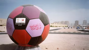 L'immagine mostra un'enorme palla da calcio decorata, posizionata in un'area all'aperto. La palla presenta colori vivaci, inclusi viola e nero, e ha delle scritte decorative, probabilmente in arabo. Sul fronte, si vede un cartello con la scritta "BIDDING NATION" e il nome "QATAR", che indica il ruolo del Qatar come nazione candidata per un evento sportivo. Sullo sfondo, si intravedono edifici moderni e strade, in un contesto urbano. L'atmosfera è illuminata da una luce chiara, suggerendo una giornata di sole.