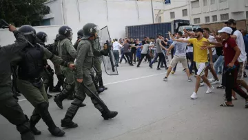 L'image montre une scène de confrontation entre des agents de sécurité en uniforme, portant des casques et des protections, et un groupe de manifestants. Les policiers avancent avec des boucliers, tandis que des personnes dans le public semblent manifester avec des gestes de défi. L'atmosphère est tendue, et le cadre urbain suggère un rassemblement intense, possiblement dans un contexte de protestation ou de manifestation. Les participants sont habillés de manière casual, et on peut voir une diversité dans leurs attitudes et leurs réactions face à la situation.
