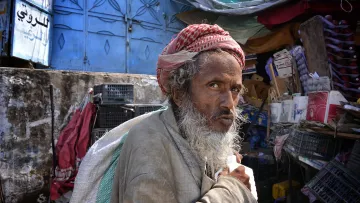 الصورة تُظهر رجلًا مسنًا، يبدو من ملامحه أنه يحمل تجاعيد تعبر عن تجارب الحياة. لديه لحية بيضاء وشعر تحت الغطاء الذي يضعه على رأسه. يرتدي ملابس بسيطة قديمة. تعكس الخلفية بساطتها وفوضاها، حيث توجد حاويات وخزائن وألوان متداخلة. تعبير وجهه يحمل مزيجًا من الحزن والتأمل، وكأنه يعيش في عالم خاص به.