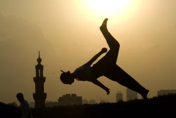 L'image montre une silhouette d'une personne effectuant un saut ou un mouvement acrobatique contre un fond de soleil couchant. À l'arrière-plan, on aperçoit une mosquée avec un minaret, ainsi que des bâtiments de la ville. L'éclairage crée un contraste fort entre la silhouette et le ciel lumineux, ajoutant une atmosphère dynamique et artistique à la scène.