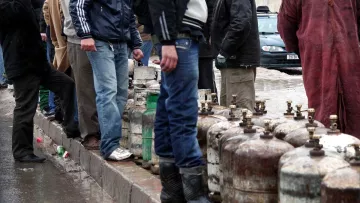 L'image montre un groupe de personnes rassemblées sous la pluie, attendant près de bouteilles de gaz. Certains portent des vêtements imperméables, comme des vestes et des manteaux, tandis que d'autres se protègent de la pluie avec des parapluies. L'atmosphère semble sombre et humide, suggérant une situation de manque ou d'attente. En arrière-plan, des voitures sont visibles, ainsi que des bâtiments. Les gens semblent engagés dans une activité collective, probablement en attendant d'acheter du gaz.