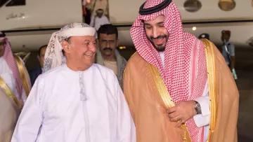 در این تصویر، دو مرد در حال خوش‌وبش با یکدیگر هستند. یکی از آن‌ها لباس سنتی عربی پوشیده و دیگری نیز لباس مشابهی به همراه عبای طلایی بر تن دارد. هر دو در حال خندیدن و گفتگو هستند و به نظر می‌رسد در یک فرودگاه یا محل نشست هستند. در پس‌زمینه، یک هواپیما دیده می‌شود و تعدادی افراد دیگر نیز در تصویر حضور دارند. جو تصویر دوستانه و گرم است.