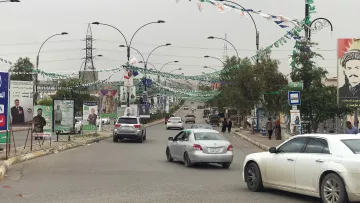 L'image montre une rue bordée de drapeaux et de bannières, probablement en préparation d'un événement ou d'une célébration. On peut voir plusieurs voitures circulant sur la route. À côté de la route, il y a des panneaux avec des affiches, ce qui indique une zone urbaine active. Des personnes se trouvent également sur le trottoir, et des arbres et des lampadaires sont présents le long de la rue, donnant un aspect dynamique à la scène. Le ciel semble nuageux, suggérant une ambiance calme.