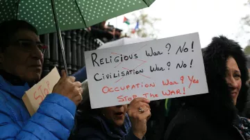 في الصورة، يظهر مجموعة من الأشخاص في تظاهرة، حيث يحمل أحدهم لافتة مكتوب عليها "الحرب الدينية؟ لا! الحرب الحضارية؟ لا! حرب الاحتلال؟ نعم! اوقفوا الحرب!"، بينما يحمل آخر مظلة خضراء. التعبيرات على وجوه المتظاهرين تدل على الإصرار والمشاركة الفعالة في الاحتجاج. الأجواء تتسم بالجدية مع خلفية غائمة أو ممطرة.