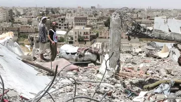 تظهر الصورة مشهدًا من مدينة مدمرة، حيث يقف رجلان على أنقاض مبنى. خلفهما، ترتفع مجموعة من المباني في المدينة، وهي تبدو متضررة بشكل كبير. يبدو أن البيئة المحيطة تعكس آثار الدمار، مع الحطام والمخلفات المنتشرة في كل مكان. السماء تبدو مشمسة، مما يبرز الف contrast بين الدمار والبيئة المحيطة.