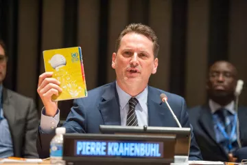 The image shows a man speaking at a podium during a conference or meeting, likely at a United Nations event. He is holding up a yellow booklet or document, which appears to be significant to his presentation. The nameplate in front of him reads "PIERRE KRAHENBUHL." In the background, there are several other individuals seated, and the setting has a formal atmosphere typical of international discussions. The man is engaged and emphasizing a point while addressing the audience.