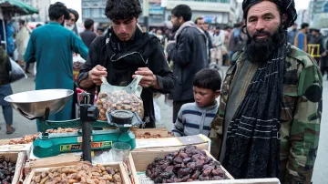 L'image montre un marché en plein air, probablement dans un pays asiatique. Au premier plan, on voit un homme portant un turban et une barbe, habillé d'une veste militaire. À côté de lui, un enfant observe. L'homme a l'air de s'occuper d'une balance et de dates, qui sont présentées dans des caisses en bois à ses côtés. En arrière-plan, on aperçoit d'autres personnes, ce qui donne une impression de foule animée et d'activité commerciale. L'environnement semble rural et traditionnel, avec des bâtiments flous au fond.