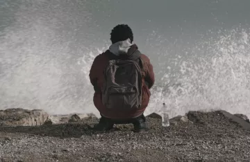 L'image montre une personne assise sur un rivage, tournant le dos à la caméra. Elle porte une veste et un sac à dos. À côté d'elle, il y a une bouteille d'eau. On peut voir des vagues qui se brisent sur les rochers, créant un effet de mouvement dans l'eau. L'atmosphère semble calme mais dynamique en raison du mouvement des vagues.