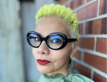 L'image montre une personne avec des cheveux très courts teints en jaune vif. Elle porte de grandes lunettes noires au style distinctif. Son maquillage comprend des lèvres rouge vif. Le fond présente un mur en briques, ce qui ajoute un contraste avec son look. Elle a une expression confiante et souriante.