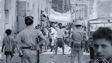 تظهر الصورة مجموعة من الأشخاص في شارع ضيق، يبدو أنهم في خضم حدث أو تظاهرة. في المقدمة، هناك رجل يرفع يديه حاملاً لافتة، بينما يتواجد أفراد من قوات الأمن بالقرب منه. يمكن رؤية الأطفال والبالغين في الخلفية، بعضهم يبدو مهتماً بالحدث. الملابس والإعداد تعكس فترة زمنية معينة، كما يظهر في التفاصيل المعمارية للمنازل. المشهد يحتوي على أجواء من الحماس والتوتر.