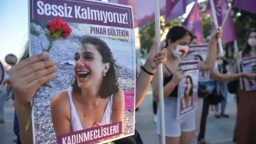L'image montre un groupe de personnes manifestant avec des pancartes. L'une des pancartes porte le visage d'une jeune femme, accompagnée d'un message en turc qui se traduit par "Nous ne restons pas silencieuses !". Une manifestante tient une fleur rouge. Les femmes portent des vêtements variés et semblent engagées dans une cause, probablement liée à la défense des droits des femmes. L'atmosphère peut être perçue comme déterminée et solidaire, avec un accent sur la mémoire d'une personne nommée Pınar Gültekin.