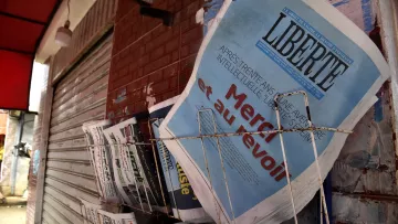 L'image montre un présentoir de journaux, où plusieurs titres sont visibles. Le journal en premier plan affiche le mot "LIBERTÉ" en lettres majuscules, accompagnés d'un sous-titre qui semble évoquer un sujet d'actualité. Les autres journaux à côté sont disposés de manière ordonnée, avec des titres variés, ce qui suggère un point de vente de presse dans un environnement urbain. Le mur en arrière-plan, ainsi que le toît rouge au-dessus, ajoutent à l'atmosphère locale de la scène.