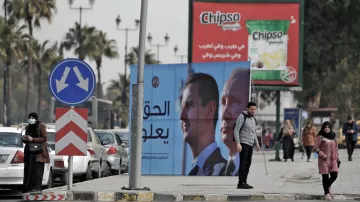 در این تصویر، خیابانی مطرح شده و در آن دو نفر در کنار پیاده‌رو ایستاده‌اند. در پس‌زمینه، چندین خودرو پارک شده و همچنین یک بیلبورد بزرگ با تصاویر و متنی به زبان عربی دیده می‌شود. این بیلبورد به احتمال زیاد به مقامات و موضوعات سیاسی اشاره دارد. تبلیغاتی برای یک برند معروف تنقلات نیز در بالای تصویر دیده می‌شود. در مجموع، تصویر حال و هوای شهری را به نمایش می‌گذارد.