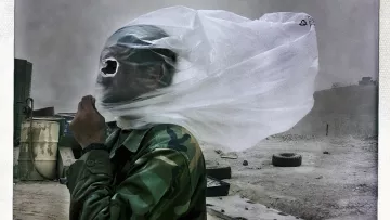 L'image montre un soldat portant un uniforme camouflé, protégé par un morceau de tissu fin qui couvre son visage. Le vent soulève le tissu, créant une atmosphère dramatique. En arrière-plan, le paysage semble poussiéreux et désertique, avec des objets flous tels que des pneus. L'ensemble de la scène évoque un contexte difficile, probablement lié à des conditions climatiques extrêmes.