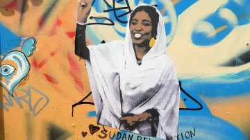 L'image montre une peinture murale colorée sur un mur. Au centre, il y a une représentation d'une femme vêtue d'une robe blanche, levant la main en signe de victoire avec deux doigts. Elle affiche un large sourire, symbolisant la positivité et la résistance. Autour d'elle, il y a des graffitis multicolores, et l'inscription "Sudan Revolution" est bien visible, ce qui indique un lien avec le mouvement révolutionnaire au Soudan. Le tout dégage une ambiance de soutien et de solidarité.