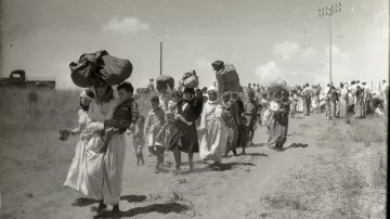 L'image montre un groupe de personnes marchant le long d'un chemin poussiéreux. Certaines femmes portent de lourdes charges sur la tête, tandis que d'autres tiennent des enfants par la main. La scène semble illustrer un moment de déplacement ou de migration, avec des paysages arides en arrière-plan. Des nuages blancs ponctuent le ciel, et on peut voir des lignes électriques au loin. L'atmosphère de la photo évoque la lutte et la détermination des personnes en marche.