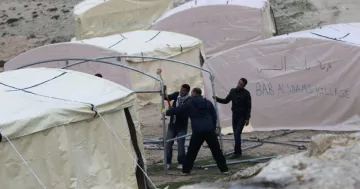 L'image montre plusieurs tentes de type campement, qui semblent être installées dans un environnement extérieur. Les tentes ont des écrits en arabe et en anglais, mentionnant "BAB AL SHAMS VILLAGE". On observe également des individus en train de travailler ensemble pour monter les structures des tentes. L'ensemble de la scène évoque des activités de construction ou d'aménagement dans un cadre probablement temporaire.