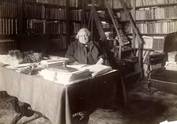 L'image montre une scène d'intérieur, probablement dans un bureau ou une bibliothèque. Un homme, apparemment âgé, est assis à une table chargée de papiers et de livres. Derrière lui, on peut voir des étagères remplies de livres. Il y a également un tapis sur le sol. Un chat, qui semble être sur la table ou à proximité, ajoute une touche de vie à l'atmosphère studieuse de la pièce. L'éclairage donne une impression de sérénité et de calme.