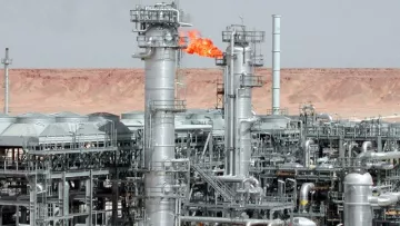 L'image montre une installation industrielle, probablement liée à l'industrie pétrolière ou gazière. On peut voir plusieurs tours métalliques, dont certaines émettent des flammes, indiquant une combustion, peut-être de gaz résiduel. L'arrière-plan présente un paysage désertique, caractérisé par des collines ou des montagnes sèches, typique des zones où se trouvent des installations énergétiques. Les structures sont en métal et semblent sophistiquées, témoignant d'une technologie avancée utilisée dans cette industrie.