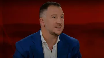 L'immagine mostra un uomo con capelli corti e una barba leggera, che indossa una giacca blu e una camicia bianca. Si trova di fronte a uno sfondo rosso. L'uomo appare in un contesto di colloquio o discussione, con un'espressione attenta e coinvolta.