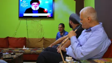 تظهر في الصورة مجموعة من الأشخاص يجلسون في غرفة معيشة مريحة، حيث يجلس رجلٌ على الأريكة وهو يدخن الشيشة. خلفه، يظهر تلفاز يُبث عليه صورة لشخص معروف يتحدث. الجدران تؤدي إلى أجواء دافئة، بينما الركن الآخر يُظهر شخصًا ضاحكًا. الألوان في المكان تتنوع بين الأخضر والبني، مما يعطي شعورًا بالراحة.
