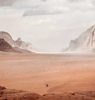 L'image montre un vaste paysage désertique. On peut voir des montagnes rocheuses au loin, entourées de dunes de sable. Le ciel est nuageux, ce qui crée une atmosphère légèrement brumeuse. Au centre, une seule silhouette humaine, probablement un cavalier, se déplace à travers ce paysage aride. La scène transmet une sensation d'immensité et de solitude.