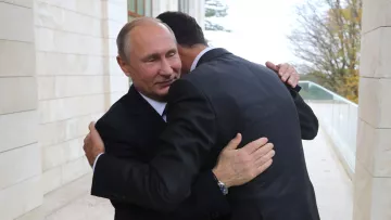 الصورة تظهر شخصين يتعانقان في مشهد يبدو وديًا. الشخص الأول يرتدي بدلة سوداء وقميصًا أبيض، بينما الشخص الثاني يرتدي بدلة داكنة أيضًا. الخلفية تظهر جزءًا من مبنى وأشجار، مما يوحي بأن اللقاء يحدث في مكان رسمي أو خارجي. تعبر تعابير الوجه عن اللحظة العاطفية لهذا اللقاء.