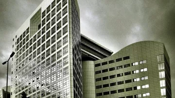 L'image montre un bâtiment moderne avec une architecture contemporaine. La structure est composée de plusieurs angles et surfaces réfléchissantes, donnant une impression de sophistication. Les vitres sont grandes, et le design présente des lignes épurées. Le ciel en arrière-plan est nuageux, ce qui peut créer une ambiance dramatique. Les tons de l'image sont en noir et blanc, accentuant les contrastes entre la lumière et l'ombre sur la façade du bâtiment.
