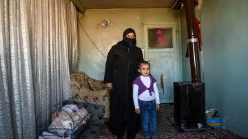 L'image montre une femme portant une tenue noire et un voile, se tenant à côté d'une petite fille. Elles sont à l'intérieur d'une pièce qui semble modeste, avec des murs verts pâles et un tapis au sol. À gauche, il y a une couchette avec un bébé endormi. Un poêle en métal est visible dans un coin de la pièce, et des rideaux en tissu léger sont tirés. L'ambiance est familiale, mais la simplicité du décor reflète une certaine précarité.