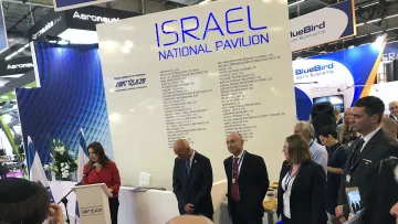The image shows a gathering at the Israel National Pavilion at an event or exhibition. A woman is speaking at a podium, while several individuals stand beside her, including a man in a suit and others who appear to be attentive to her speech. The background features a large display with the words “ISRAEL NATIONAL PAVILION” and various company names listed. The atmosphere seems formal, with an audience present, possibly indicating a business or governmental event.