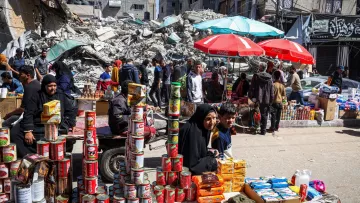 L'immagine mostra una scena di un mercato affollato, situato tra rovine di edifici. In primo piano, un gruppo di persone sta vendendo prodotti alimentari, con diversi ripiani di scatole e cibo in mostra. Sono visibili delle donne e dei bambini, alcuni seduti e altri in piedi, mentre diversi ombrelloni rossi forniscono ombra. Sullo sfondo, ci sono detriti di edifici crollati, che testimoniano una situazione di crisi. L'atmosfera sembra vivace nonostante le difficoltà evidenti.