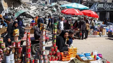 L'image montre une scène d'un marché en plein air, où des gens se déplacent parmi des étals de produits. Au premier plan, une femme en hijab et un enfant sont assis devant une pile de conserves et d'autres produits alimentaires. En arrière-plan, des bâtiments en ruines et des débris sont visibles, suggérant un contexte de destruction. Des parasols de différentes couleurs offrent de l'ombre aux vendeurs et aux clients. L'atmosphère semble chargée de vie malgré le décor dévasté.