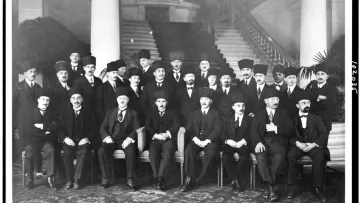 L'image présente un groupe d'hommes, probablement d'importance politique ou officielle, assis et debout dans un cadre formel. Ils portent des costumes sombres et des chapeaux traditionnels, et certains ont des moustaches. La scène se déroule dans un intérieur élégant, avec des colonnes décoratives et des plantes en pot en arrière-plan. L'atmosphère dégage une impression de solennité et de cérémonie, suggérant un événement ou une réunion importante.