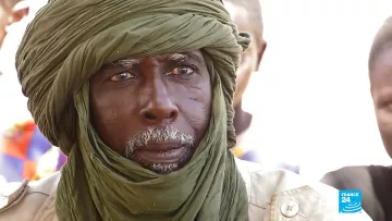 The image features an elderly man with a distinguished appearance. He is wearing a green turban wrapped around his head, which adds to his dignified look. His face shows signs of age, with deep wrinkles and a white beard, suggesting wisdom and experience. The background appears to be a social setting with other individuals present, though they are not clearly defined in the image. The focus is on the man, capturing a solemn expression that hints at deep thought or contemplation.