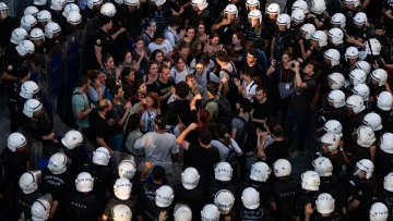 L'image montre une scène de manifestation où une foule de personnes se trouve face à des policiers en tenue anti-émeute. Les manifestants semblent discuter ou protester, tandis que les policiers forment une ligne serrée. L'atmosphère est tendue, et une partie de la foule semble concentrée sur un point, indiquant une interaction ou un affrontement verbal. Les casques des policiers et leur équipement renforcent l'impression de confrontation.