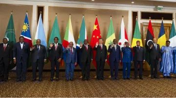 L'image montre un groupe de personnes debout en rangée, saluant. Elles se trouvent devant une série de drapeaux représentant différents pays. Les personnes portent des costumes formels et semblent participer à un événement diplomatique ou à un sommet international. Les drapeaux en arrière-plan incluent ceux de l'Union africaine, de la Chine, ainsi que d'autres nations. L'ambiance générale semble conviviale et respectueuse.