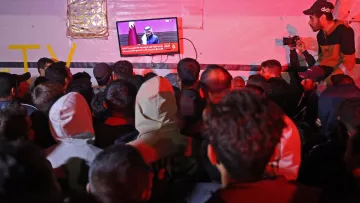 L'immagine mostra un gruppo di persone affollate intorno a un televisore. Sullo schermo è visibile un uomo che parla, mentre la parte inferiore dello schermo mostra un testo in arabo. L'illuminazione dell'ambiente è scarsa e i partecipanti indossano vari tipi di abbigliamento, inclusi cappucci e giacche. Alcuni presenti sono distratti dal loro smartphone o sembrano concentrati su ciò che viene trasmesso in TV.