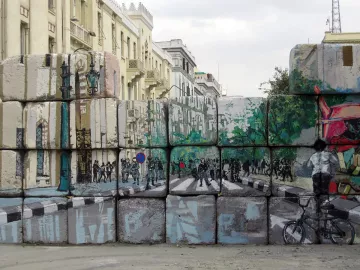 L'image montre un mur de blocs de béton, sur lequel sont peints des paysages urbains. La peinture représente une rue avec des arbres, des piétons et des cyclistes. On peut voir des silhouettes de personnes et des éléments de la ville, comme des lampadaires. Ce contraste entre le mur brut et l'art coloré apporte une touche dynamique à l'environnement urbain. Le ciel est nuageux, ajoutant une atmosphère un peu dramatique à la scène.