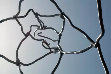 L'image montre une structure artistique composée de fils métalliques entrelacés. Les fils sont courbés et forment des nœuds à certains endroits, créant un motif complexe et abstrait. On peut voir le ciel en arrière-plan, ce qui met en valeur les formes des fils. L'ensemble dégage une impression de légèreté et de fluidité, tout en étant à la fois structurée et organique.