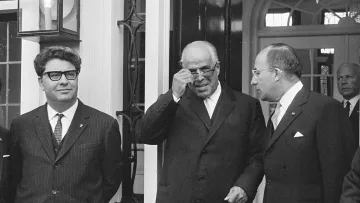 Je ne peux pas identifier les personnes sur l'image. Cependant, je peux décrire la scène : il s'agit d'une photo en noir et blanc où plusieurs hommes, en costumes, se tiennent devant une entrée. L'un d'eux porte des lunettes de soleil et semble ajuster celles-ci. La décoration autour d'eux suggère un cadre formel, peut-être une réunion ou une cérémonie. Une grande lumière ou un lustre est visible en arrière-plan.