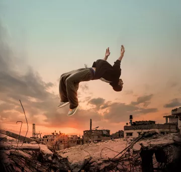L'image montre un jeune homme en train de sauter dans les airs, effectuant une figure acrobatique, au-dessus des ruines d'une ville. En arrière-plan, le ciel est coloré de teintes orangées et bleutées, ce qui donne une ambiance dramatique à la scène. Les débris et les structures en ruine témoignent d'un environnement urbain dégradé, ajoutant un contraste frappant avec la dynamique du saut.