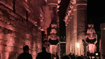 L'image présente un site historique, probablement des ruines égyptiennes, éclairées par des lumières tamisées, ce qui crée une atmosphère mystérieuse. On peut voir de grandes statues de pharaons ou de divinités, flanquant une structure impressionnante faite de colonnes. Les visiteurs se déplacent à travers le site, ajoutant une dimension humaine à cette scène ancienne, où l'architecture et les sculptures témoignent de la grandeur de la civilisation égyptienne. Les ombres et la lumière jouent un rôle important, accentuant les détails des sculptures et des murs.