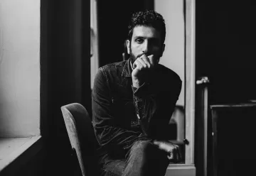 L'image montre un homme assis sur une chaise dans un intérieur sombre. Il a une barbe et porte une chemise en denim. Son expression est réfléchie, avec une main sur le menton, tandis qu'il regarde en direction de la caméra. L'éclairage met en valeur les contours de son visage et crée une ambiance introspective. Des éléments de l'environnement, comme une fenêtre et un miroir, ajoutent de la profondeur à la composition.