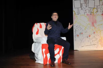 The image features a man sitting on a uniquely designed chair that has a colorful, artistic pattern, predominantly featuring red and white. He is wearing a dark outfit, including a jacket and glasses, and appears to be expressing himself with his hands raised, as if making a point during a performance or presentation. The background includes a large, stylized backdrop with a simple, colorful design. The setting seems to be a stage, suggesting a theatrical or artistic context.