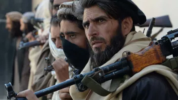 The image features a group of individuals standing closely together, each holding firearms. They appear to be dressed in traditional clothing, with some wearing headscarves and others with their faces partially obscured. The focus is on one man in the foreground, who has a serious expression and a well-groomed beard. The background is unfocused, but it suggests a rugged, possibly desert-like environment. The overall mood conveys tension or readiness.