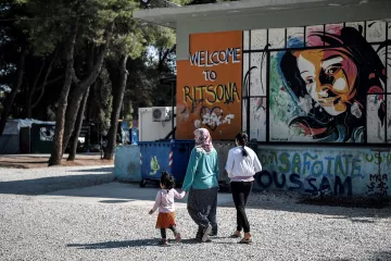 L'image montre un groupe de personnes marchant sur un chemin en gravier. À gauche, on peut voir un conteneur à ordures et à droite, un mur peint avec des graffitis colorés, dont le mot "Welcome to Ritsona" et le portrait d'une fille. Les personnes présentes semblent être une mère accompagnée de deux enfants, l'un d'eux tenant la main de la femme. L'environnement végétal suggère un cadre tranquille, malgré la situation.
